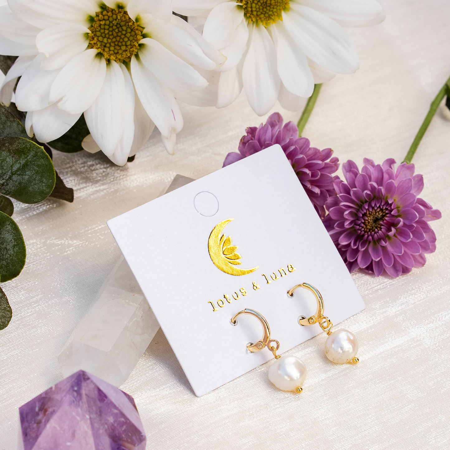 Gold earrings with pearls on a branded card surrounded by flowers and crystals