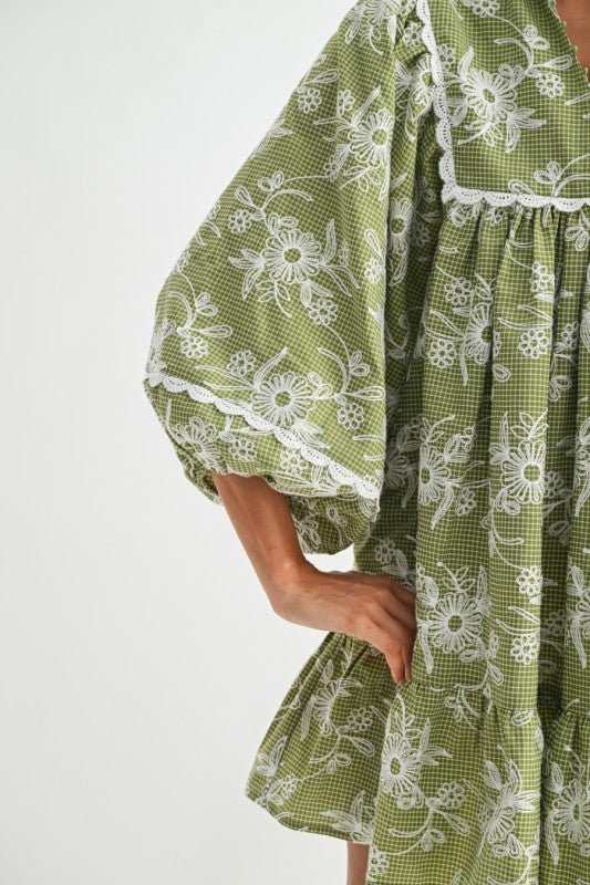 Close-up detail of green gingham dress with white embroidered flowers and scalloped lace edges on balloon sleeves — soft cotton cottagecore fabric texture.