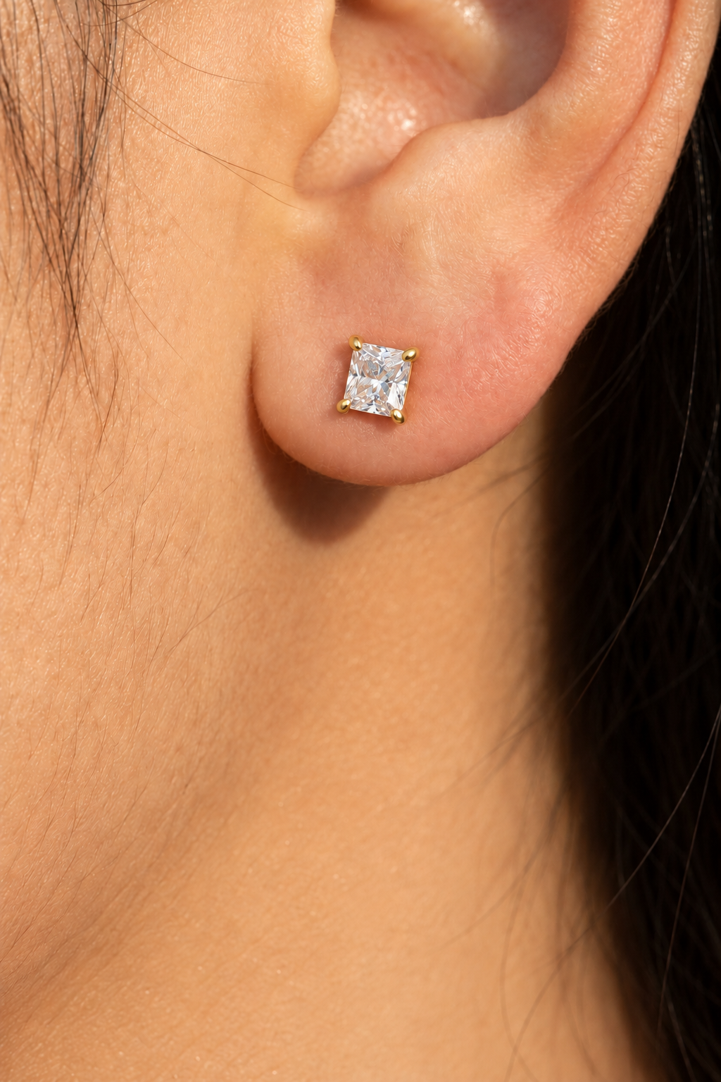 A close-up of a person's ear wearing a small square stud earring with a clear gemstone in a gold setting.