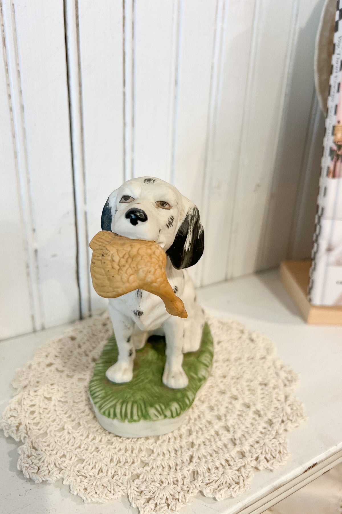 A ceramic figurine of a white and black spotted dog sitting on a green base, holding a golden-brown bone in its mouth, displayed on a cream lace doily against a white paneled background.