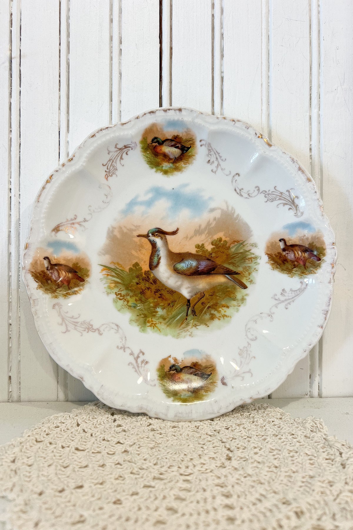 A vintage ceramic plate with a scalloped edge, featuring hand-painted duck illustrations in a natural landscape. The plate is displayed against a white wooden paneled background on a textured cream crochet doily.