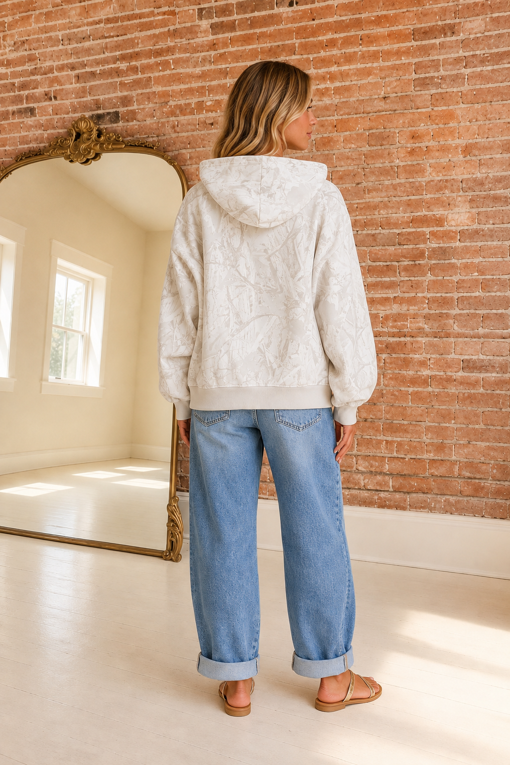 A woman stands with her back to the camera wearing a light camouflage hooded sweatshirt, light-wash blue jeans, and tan sandals in a room with a brick wall and a large mirror.