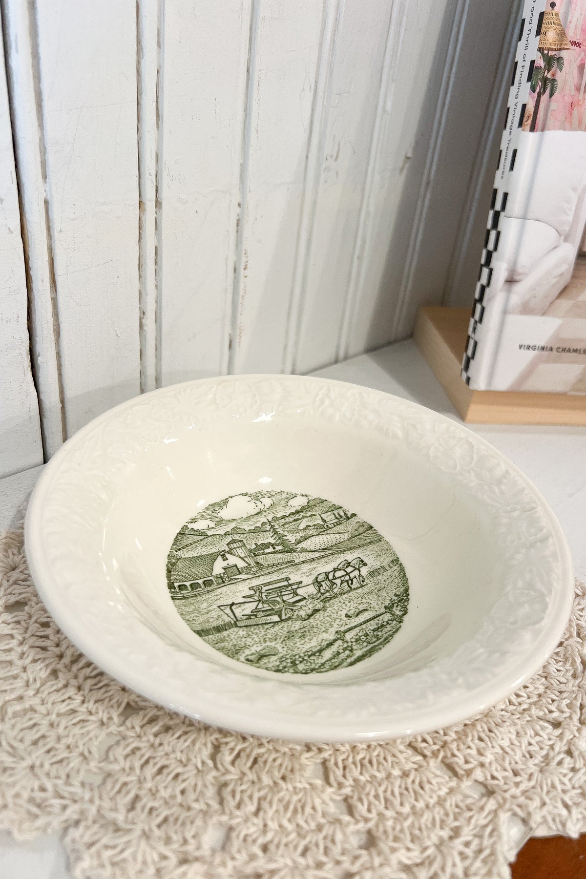 An off-white ceramic bowl with embossed floral detailing around the rim and a green vintage landscape illustration printed in the center, resting on a cream lace doily against a white paneled wall.
