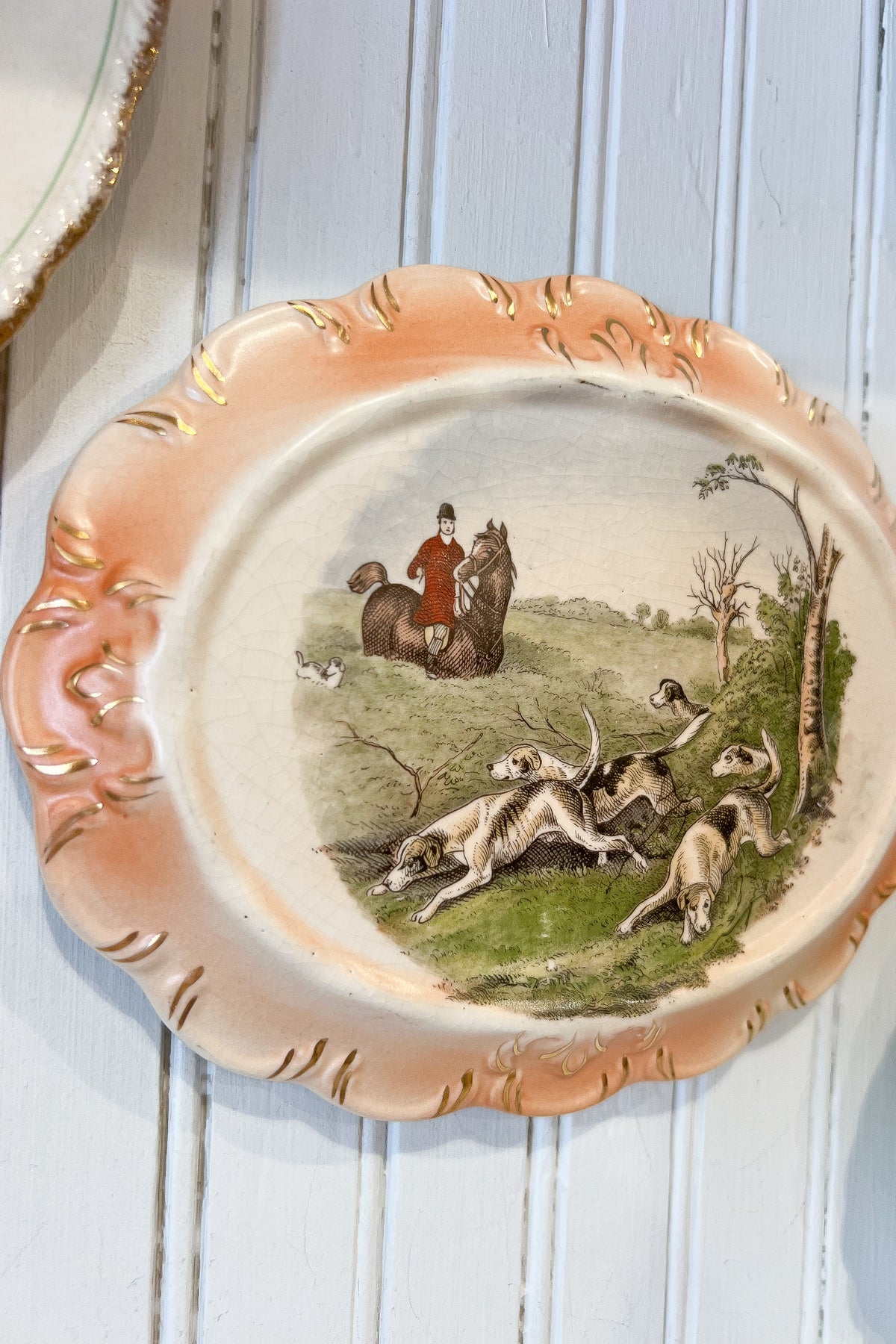 An oval decorative ceramic plate with a scalloped rim, featuring a hand-painted hunting scene with hounds, a horse, and a rider. The plate has a peach and gold rim with a cream center, displayed against a white painted wood paneled wall.