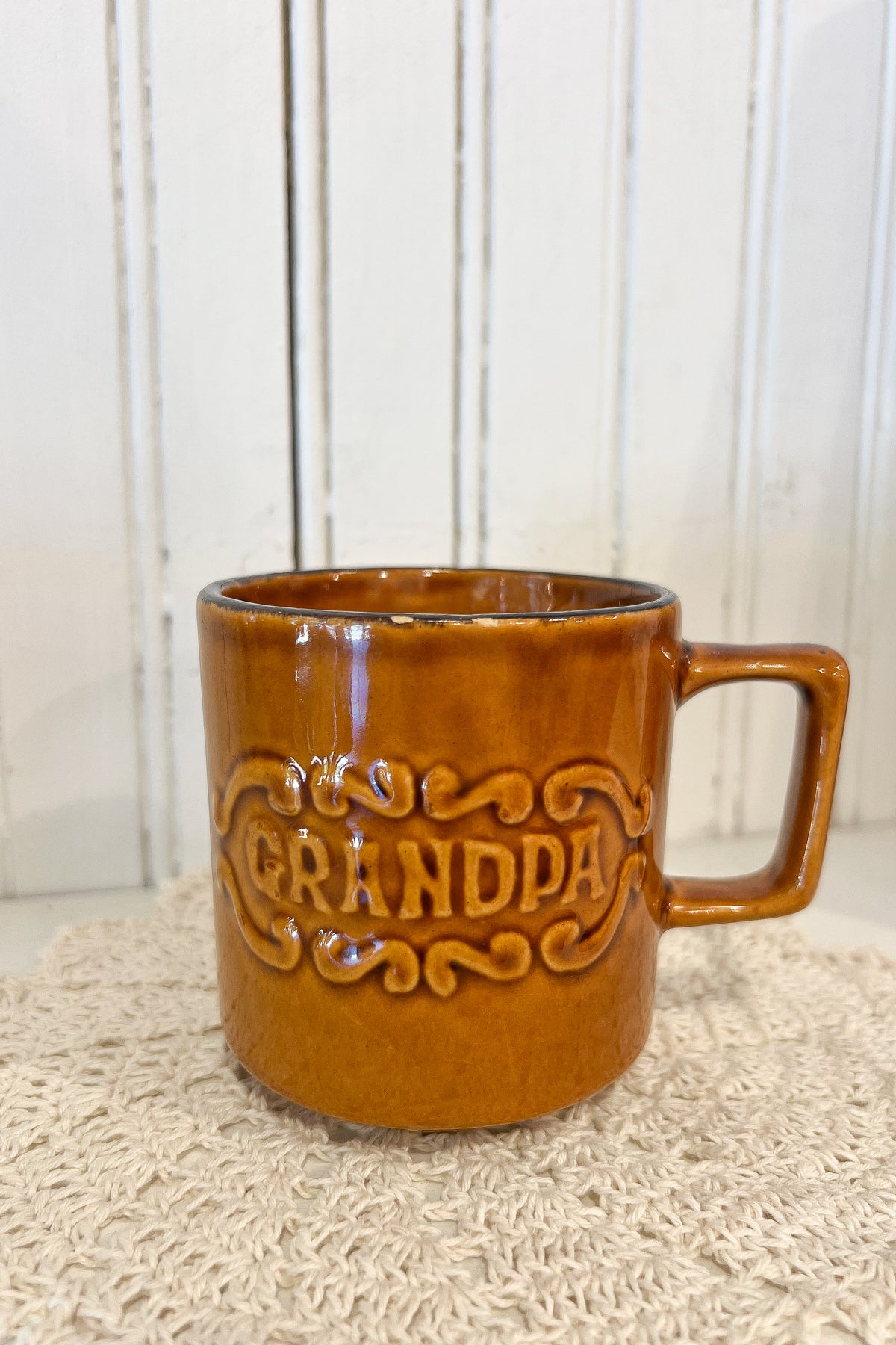 An amber-brown glazed ceramic mug with a handle, featuring an embossed 'GRANDPA' design on the front, resting on a textured beige woven placemat against a white paneled background.