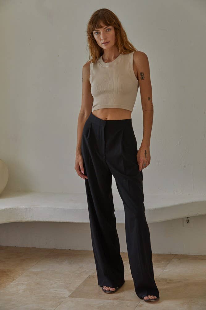 Woman wearing a beige sleeveless top and black wide-leg pants against a plain wall.