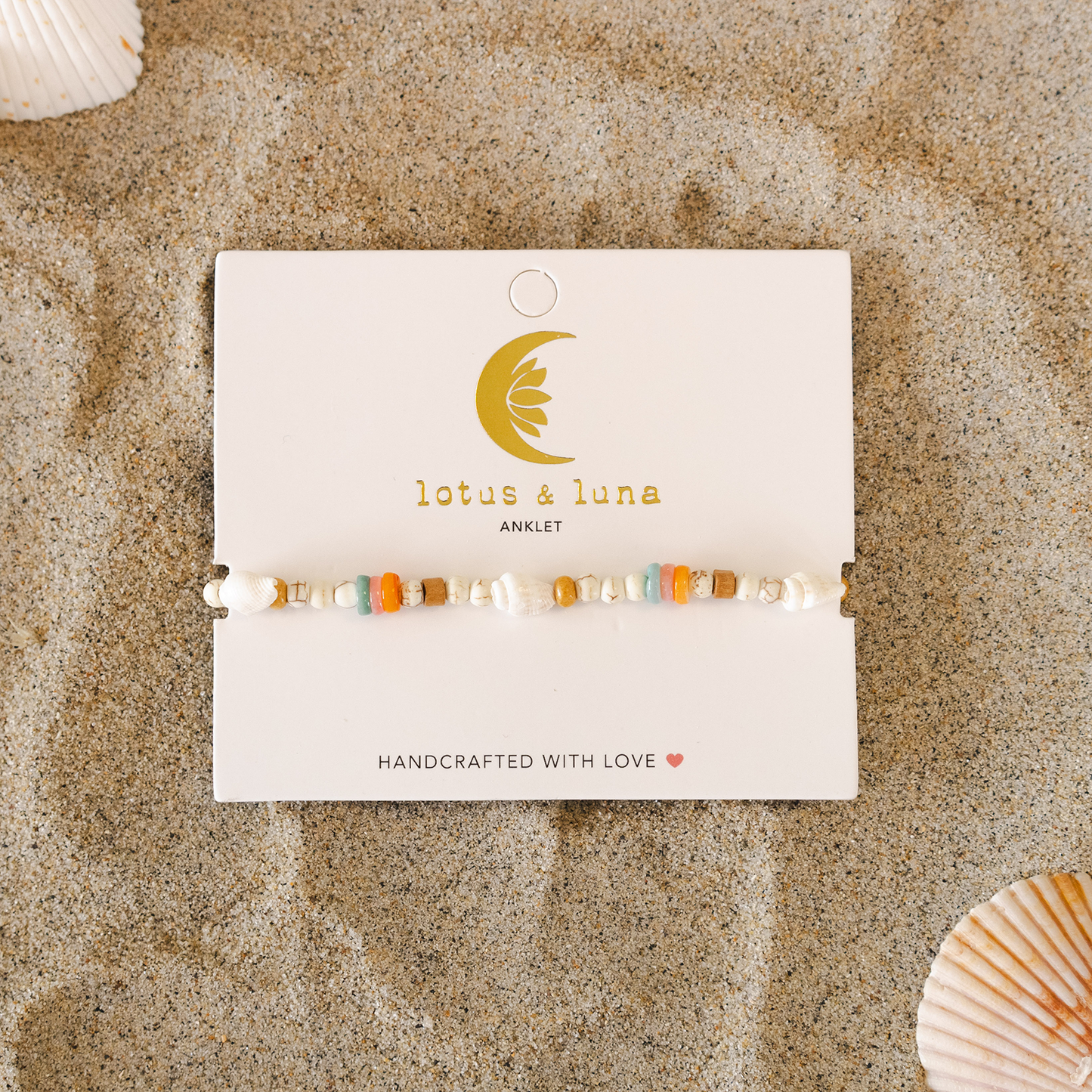 Beaded anklet on a 'lotus & luna' card with seashells around on sand