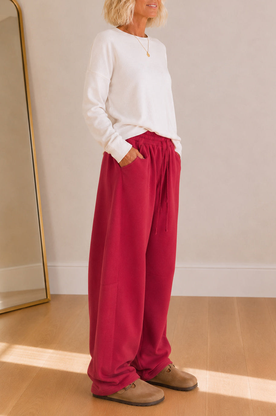 A woman wearing a white long-sleeve top and bright magenta wide-leg pants, standing in a brightly lit room with a wooden floor and a gold-framed mirror.