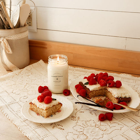 Lit Raspberry Vanilla soy candle in farmhouse kitchen setting with raspberry cake and fresh berries.