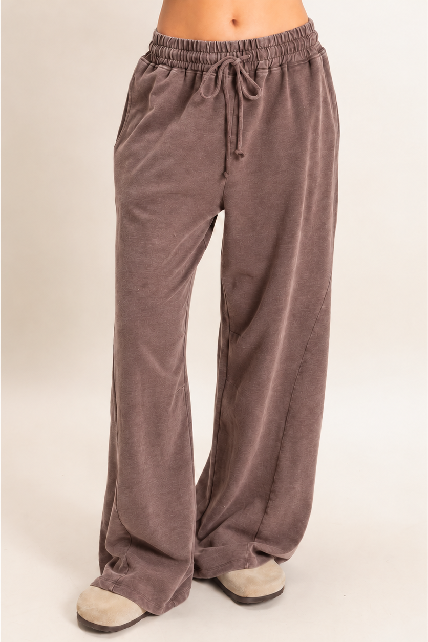 A person wearing loose, wide-leg lounge pants in a muted taupe-brown color, paired with light-colored slip-on shoes, against a plain light beige studio background.