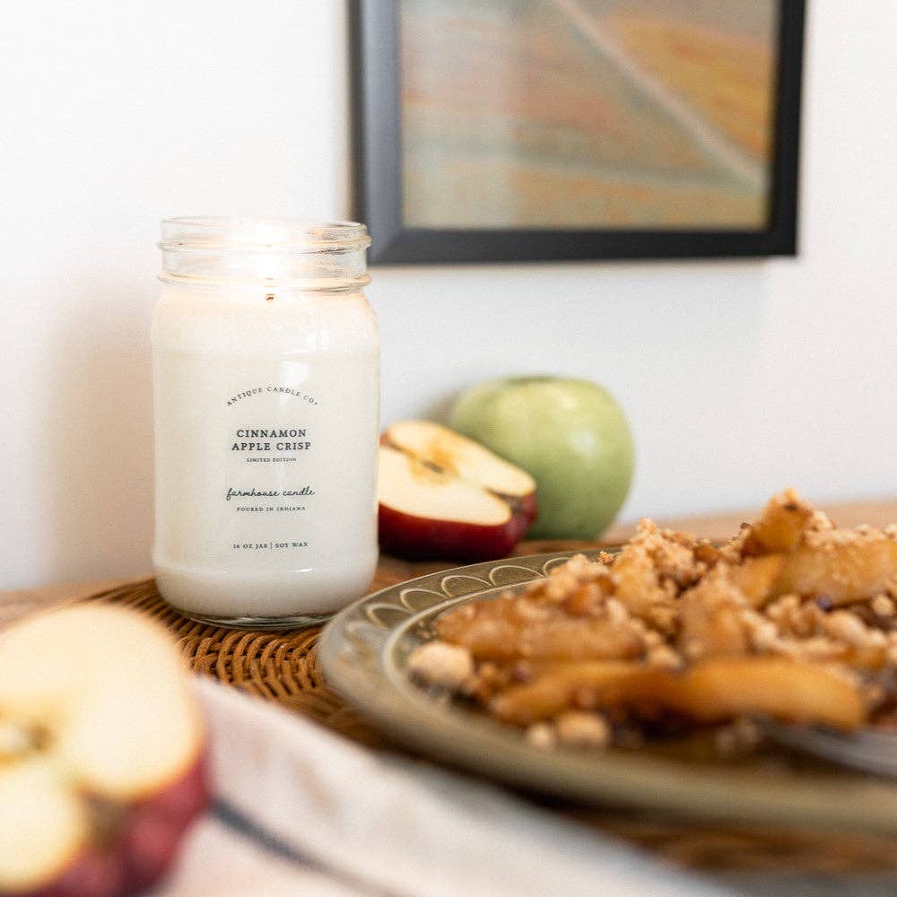 Candle labeled 'Cinnamon Apple Crisp' on a table with apples and a plate of dessert.
