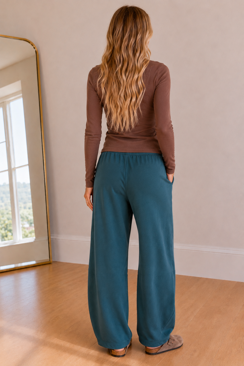 A woman with long blonde wavy hair is shown from behind wearing a brown long-sleeve top and teal wide-leg pants in a brightly lit room with a wooden floor and a mirror.