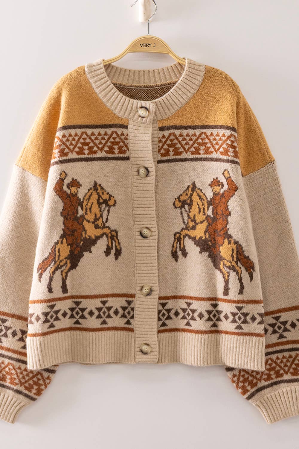 A cream and mustard yellow knit cardigan sweater with brown geometric patterns and cowboy-on-buckaroo designs, hanging on a wooden hanger against a plain white wall.