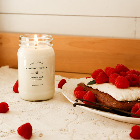 Raspberry Vanilla 16 oz soy candle by Antique Candle Co lit beside raspberry dessert with fresh berries and vanilla beans.
