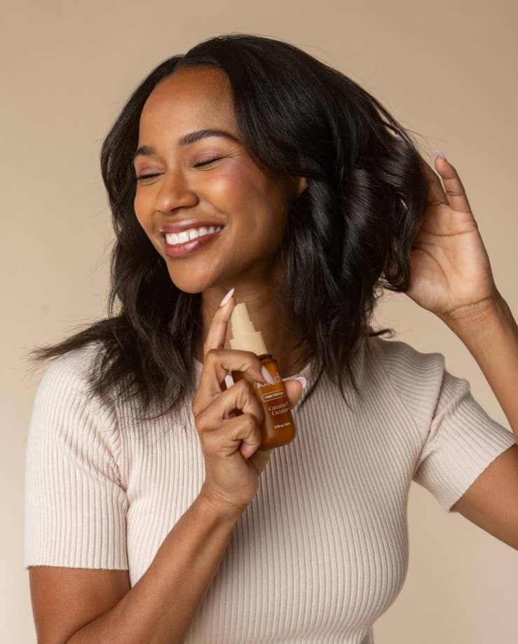 A woman with dark, wavy hair smiling while holding a bottle of hair product and spraying it. She is wearing a beige ribbed short-sleeve top against a solid beige background.