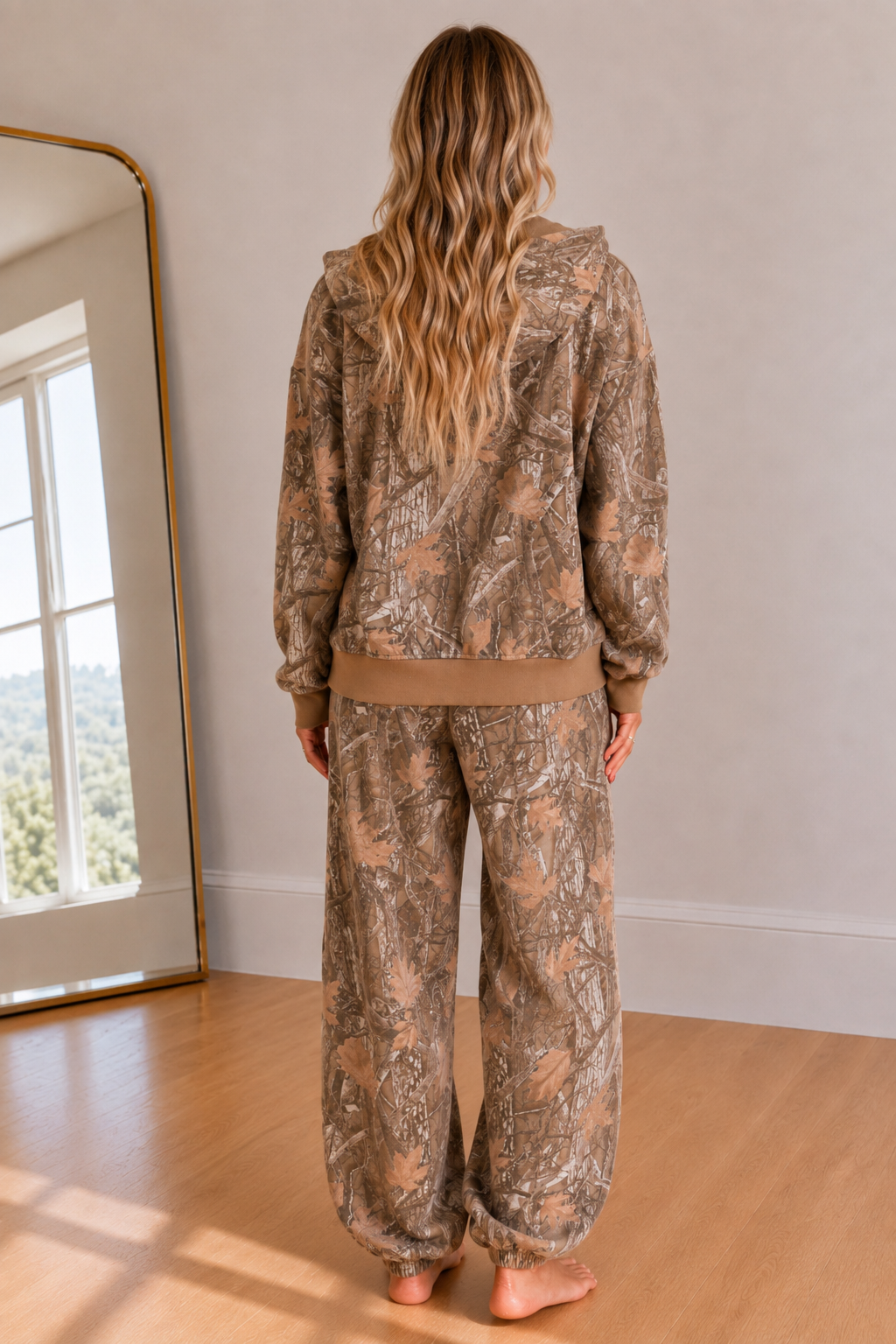 A woman stands with her back to the camera wearing a matching camouflage hoodie and sweatpants set in a bright room with a wooden floor and a large mirror.