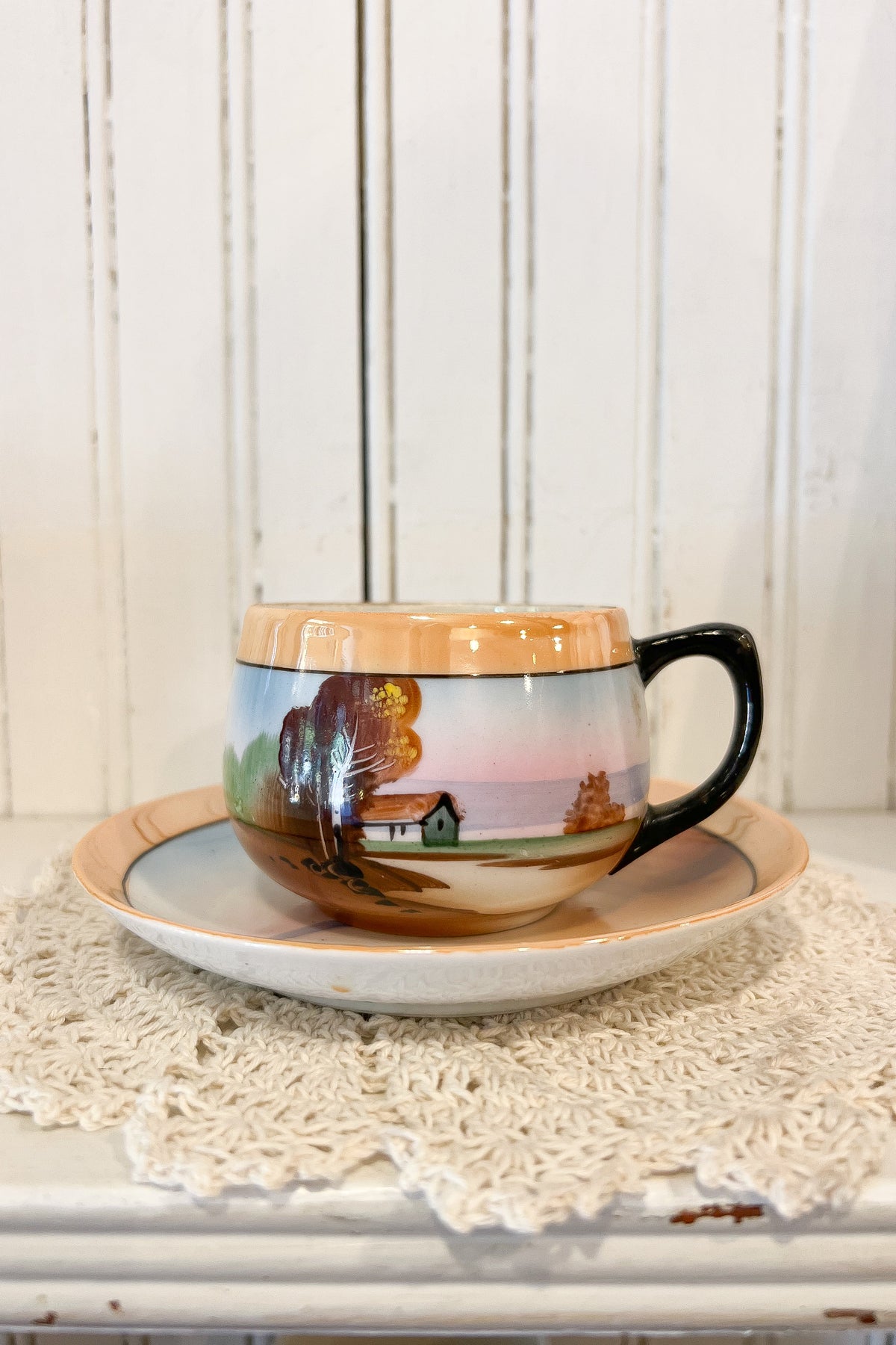 A ceramic coffee mug with a hand-painted autumn landscape scene, resting on a matching saucer over a white lace doily against a white wood-paneled background.