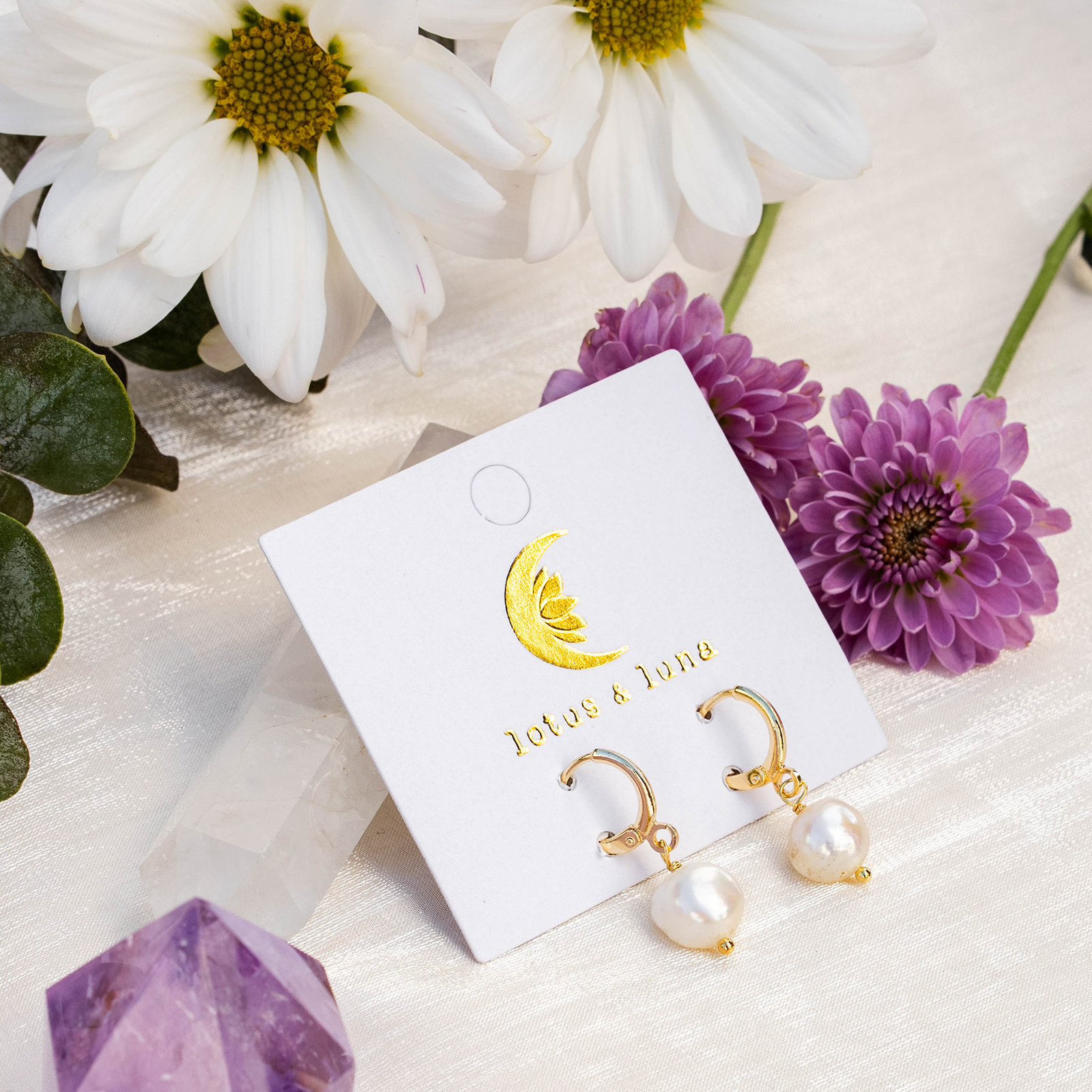 Gold earrings with pearls on a branded card surrounded by flowers and crystals