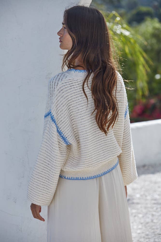 Woman wearing a light-colored knitted cardigan with blue accents against a white wall.