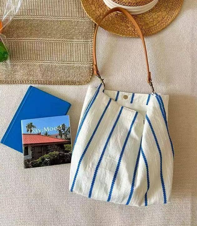 Ivory woven shoulder bag with cobalt blue stripes, tan shoulder strap, and relaxed slouchy silhouette.