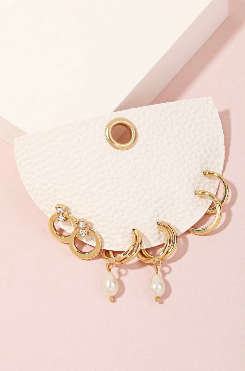 Gold earring set displayed on a textured white card against a soft pink background, featuring crystal hoops, gold hoops, and pearl drop earrings — feminine, elegant, gift-ready jewelry.