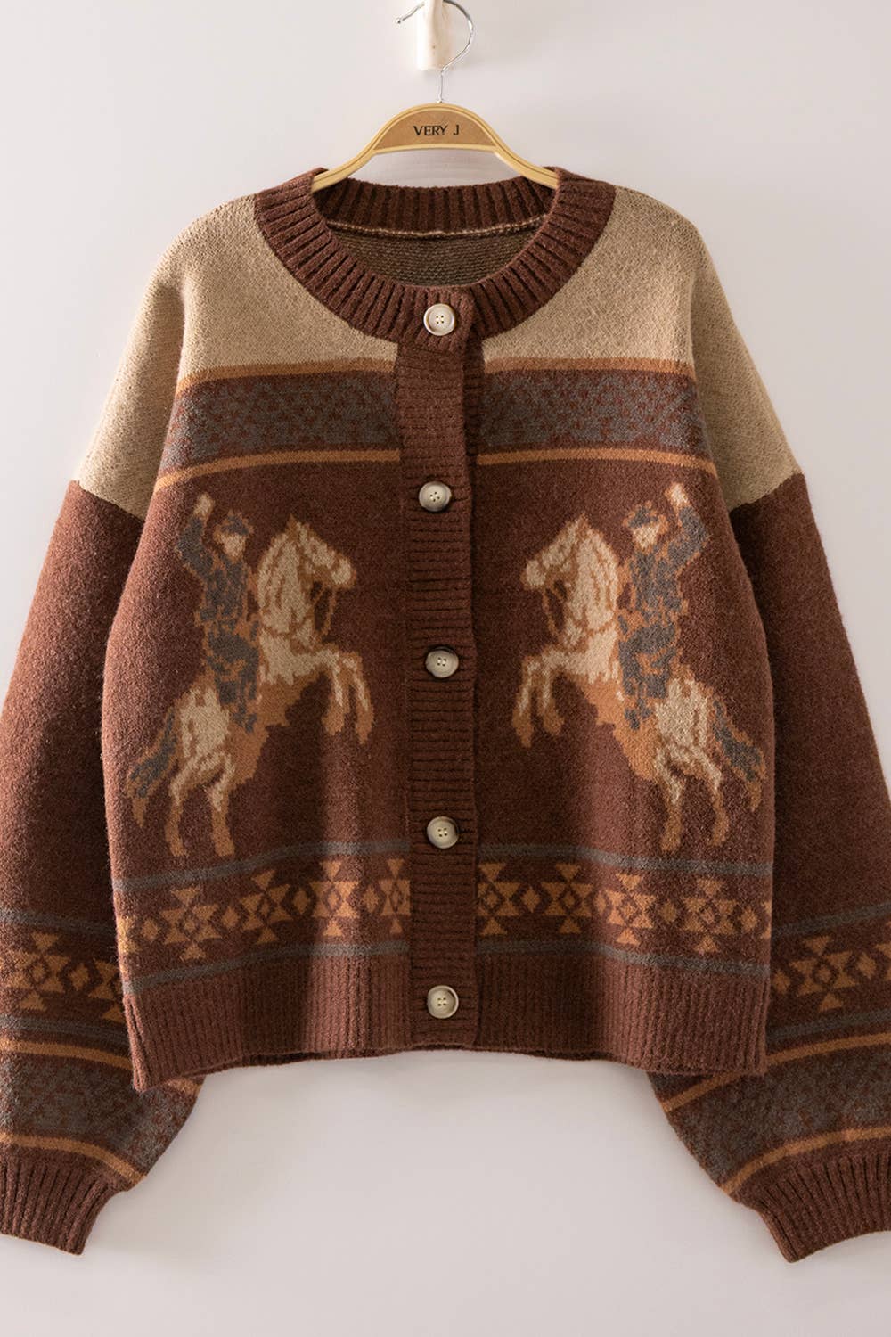 A brown and beige knit cardigan sweater with a western theme, featuring two cowboy riding horse motifs on the front. The cardigan has a ribbed crew neck, button-up front with silver buttons, and patterned bands across the chest and sleeves, hanging on a wooden hanger against a plain white wall.
