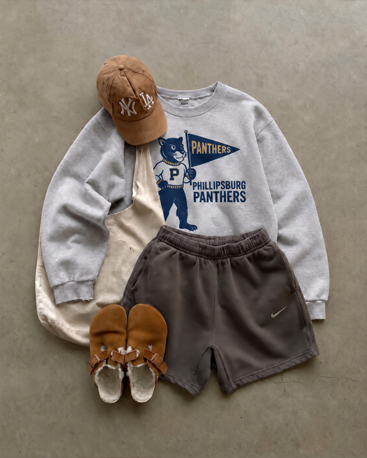 Ash grey crewneck sweatshirt styled with a tan cap, dark shorts, and sandals. The front features a vintage-inspired Phillipsburg Panthers mascot holding a navy pennant with gold lettering.