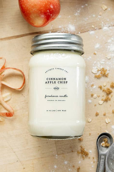 A jar candle labeled 'Cinnamon Apple Crisp' filled with creamy off-white wax and topped with a silver metal lid, styled on a light wood surface with apple slices, cinnamon sticks, oats, and metal spoons.