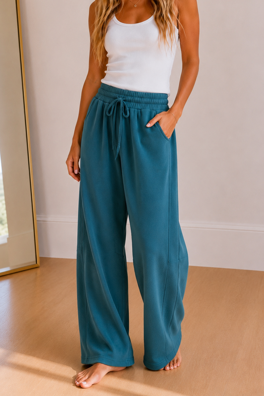 A woman stands barefoot in a bright room wearing loose teal drawstring pants with a white tank top. A tall floor mirror with a gold frame is visible on the left side of the room.