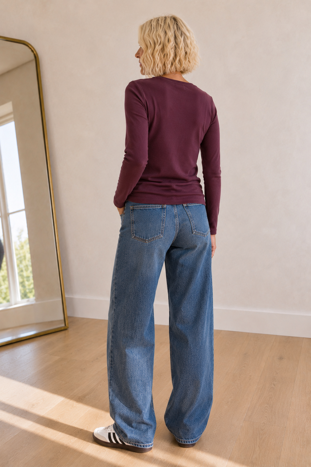 A woman with blonde hair is shown from behind wearing a maroon long-sleeve top and wide-leg blue jeans, standing in a room with light wood floors and a mirror on the left.