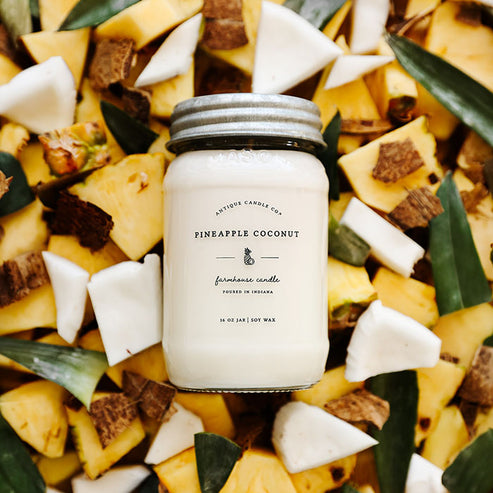 Pineapple Coconut 16 oz soy candle surrounded by fresh pineapple chunks and coconut pieces showcasing tropical summer scent.
