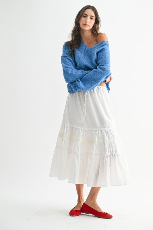 Woman wearing a slouchy blue knit sweater off one shoulder with long ribbed cuffs, styled casually with a white drawstring skirt.