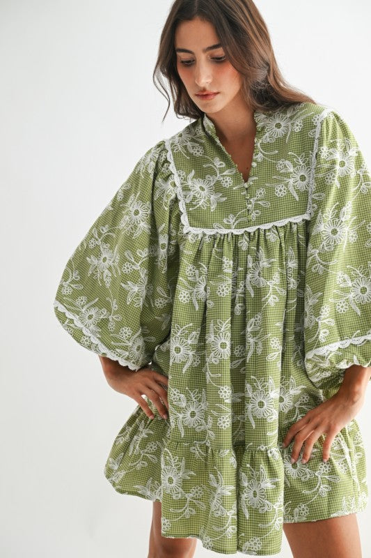 Model wearing an olive green gingham mini dress with white floral embroidery and lace trim, featuring voluminous puff sleeves and a tiered skirt — cottagecore boho style.