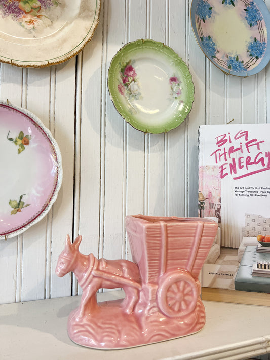 Vintage pink ceramic donkey planter with wagon detail styled on a cottage shelf with floral wall plates and nostalgic decor.