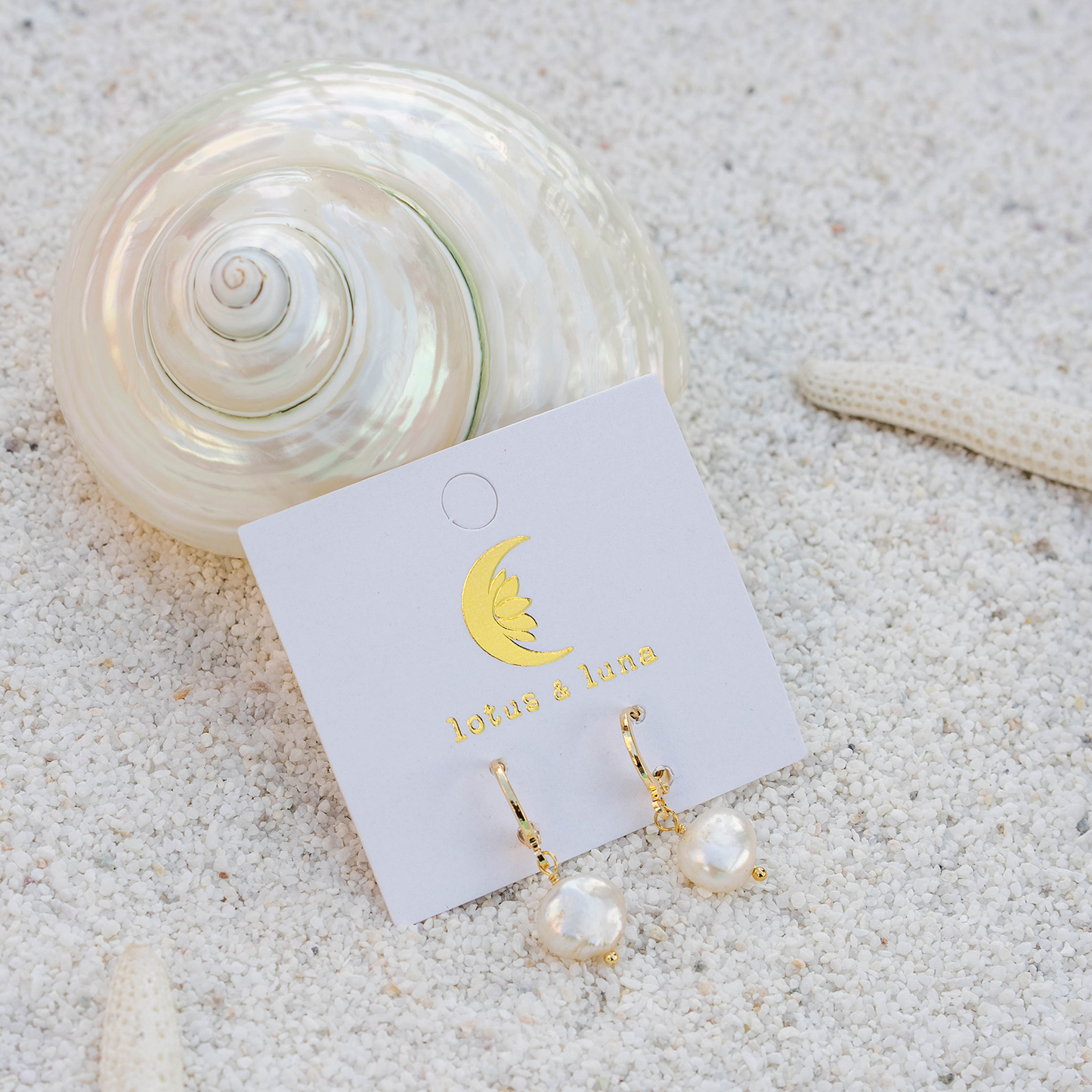 Gold and pearl earrings on a card with a shell and starfish on sand