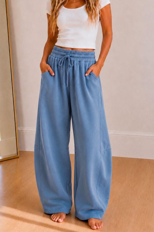 A person wearing wide-leg dusty blue lounge pants with an elastic drawstring waist and side pockets, paired with a white cropped tank top, standing barefoot on a light wood floor.
