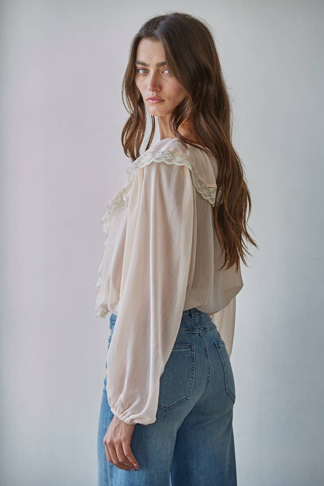 Woman wearing a beige blouse with lace details and blue jeans against a plain background