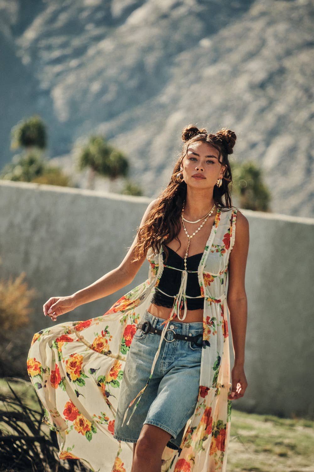 Woman in a floral dress and denim shorts standing in a desert landscape with mountains in the background.
