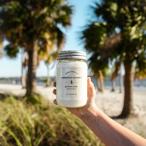 Pineapple Coconut soy candle held at beach with palm trees and ocean backdrop for tropical summer home fragrance.