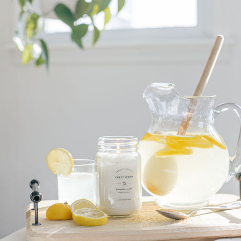 Sweet Lemon soy candle styled beside fresh lemonade pitcher and sliced lemons in bright sunlit kitchen.