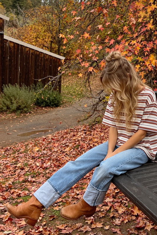 Model wearing an ivory and rust brown striped relaxed-fit tee with light-wash jeans and brown boots among colorful autumn leaves.