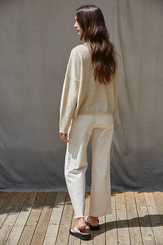 Woman wearing a beige sweater and white pants standing on a wooden floor with a gray backdrop