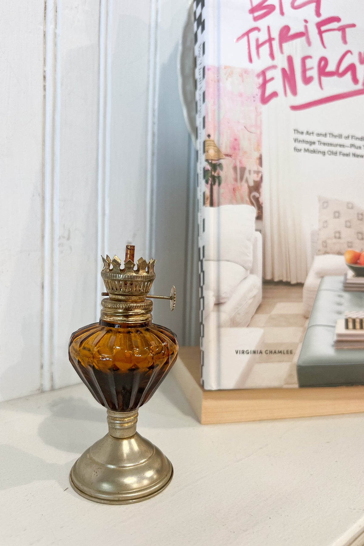 A vintage-style amber glass oil lamp with a brass-toned base and crown cap sits on a white surface beside a book titled "Big Theft Energy" against a white paneled wall.