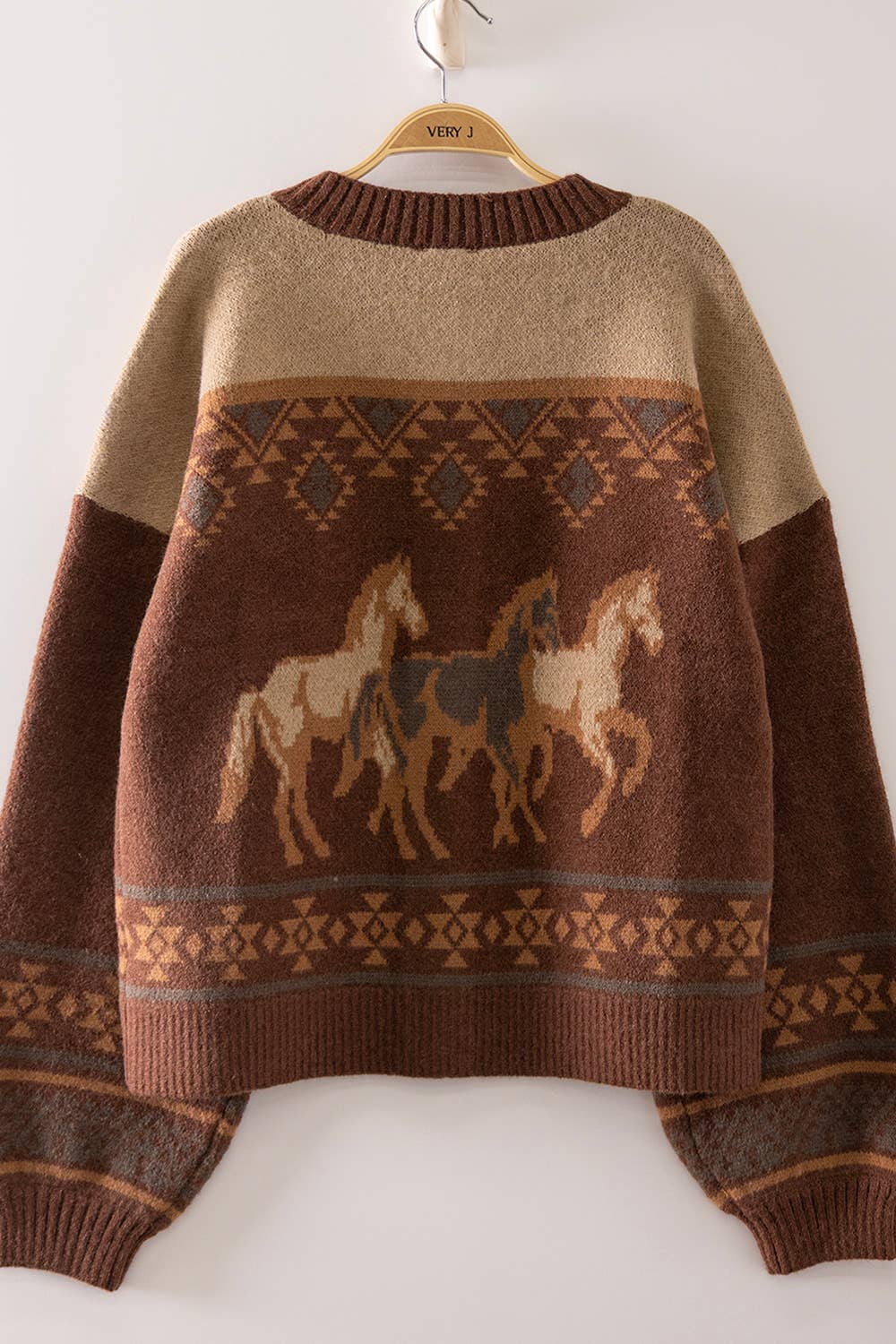 A brown and beige knit sweater with a geometric pattern across the chest and three stylized horse figures, hanging on a wooden hanger against a plain white wall.