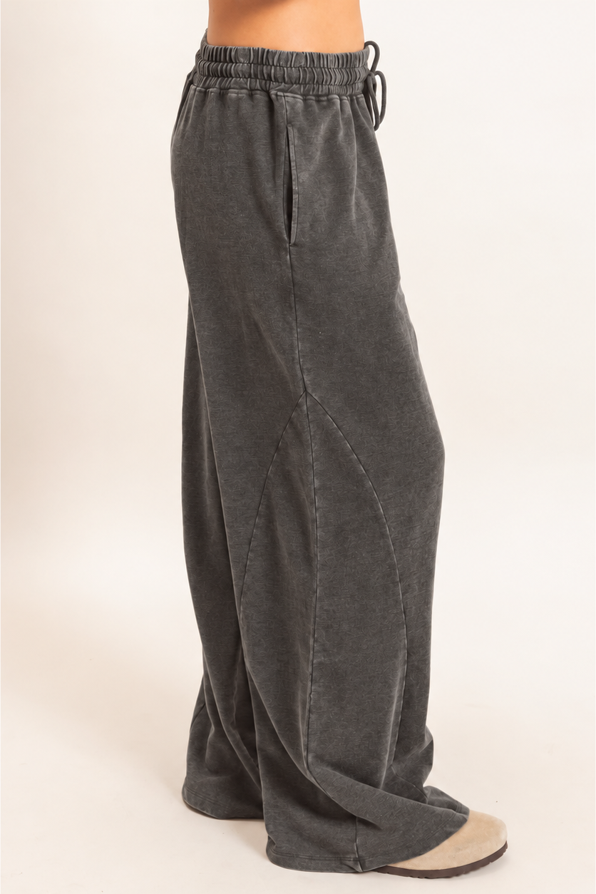 A person wearing dark charcoal gray wide-leg sweatpants with an elastic drawstring waist, shown from the waist down against a plain light beige studio background.