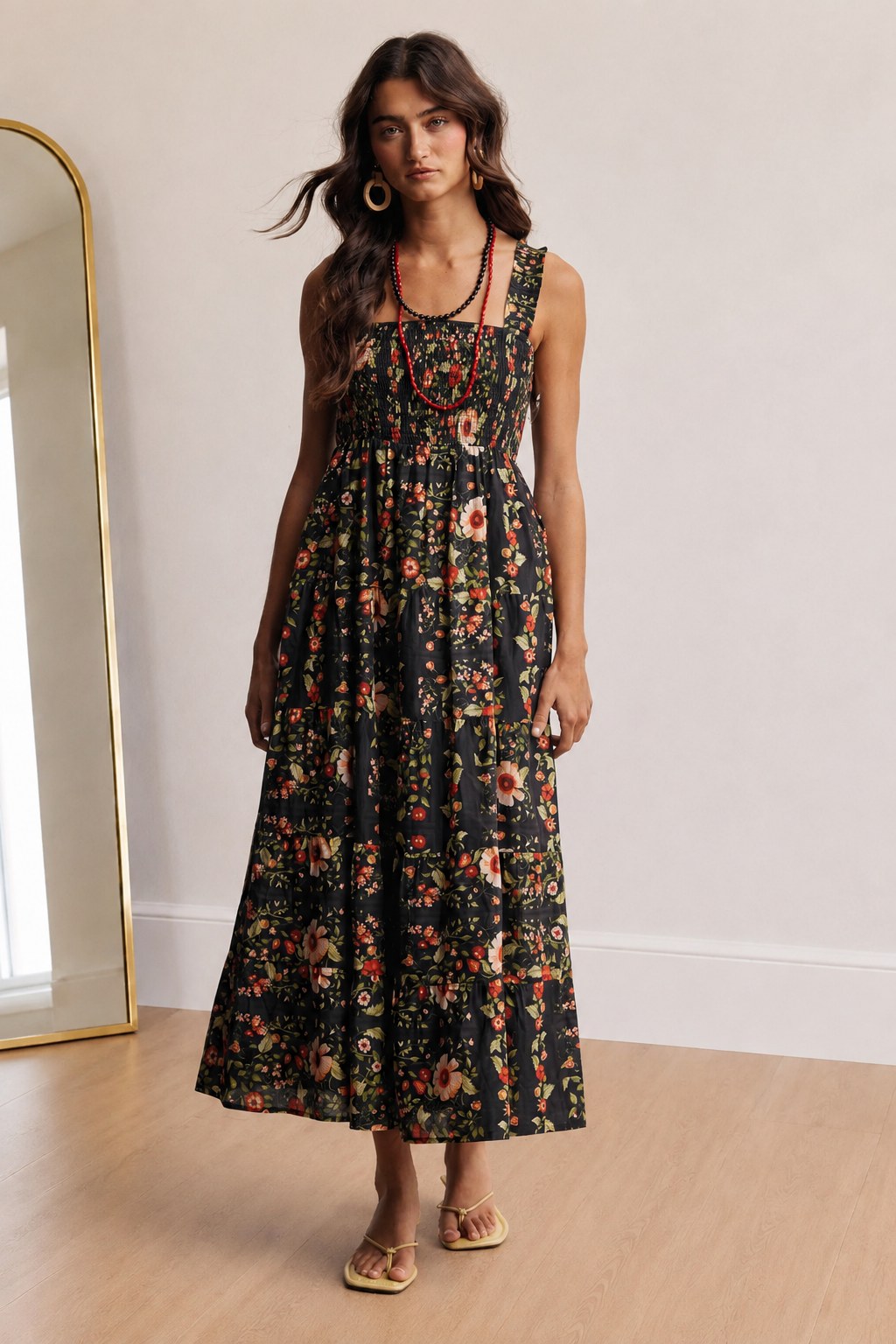 A woman models a sleeveless black maxi dress with a dense red, orange, and yellow floral print. She is standing in a brightly lit room with light wood floors and a white wall, next to a gold-framed mirror.