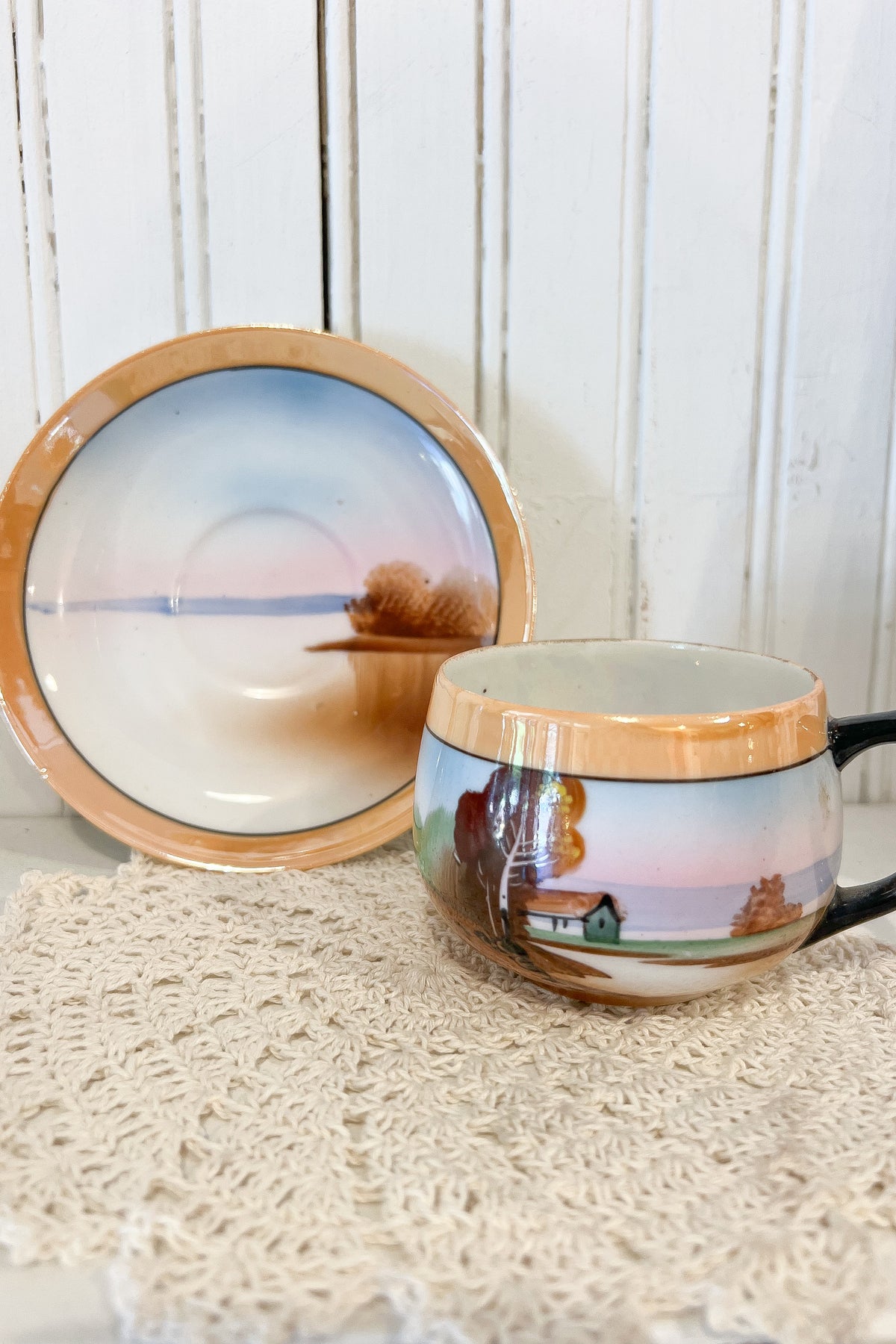 A ceramic cup and matching saucer featuring a hand-painted autumn landscape scene with a small house, trees, and a soft pastel sky, displayed on a cream lace doily against a white wood paneled background.