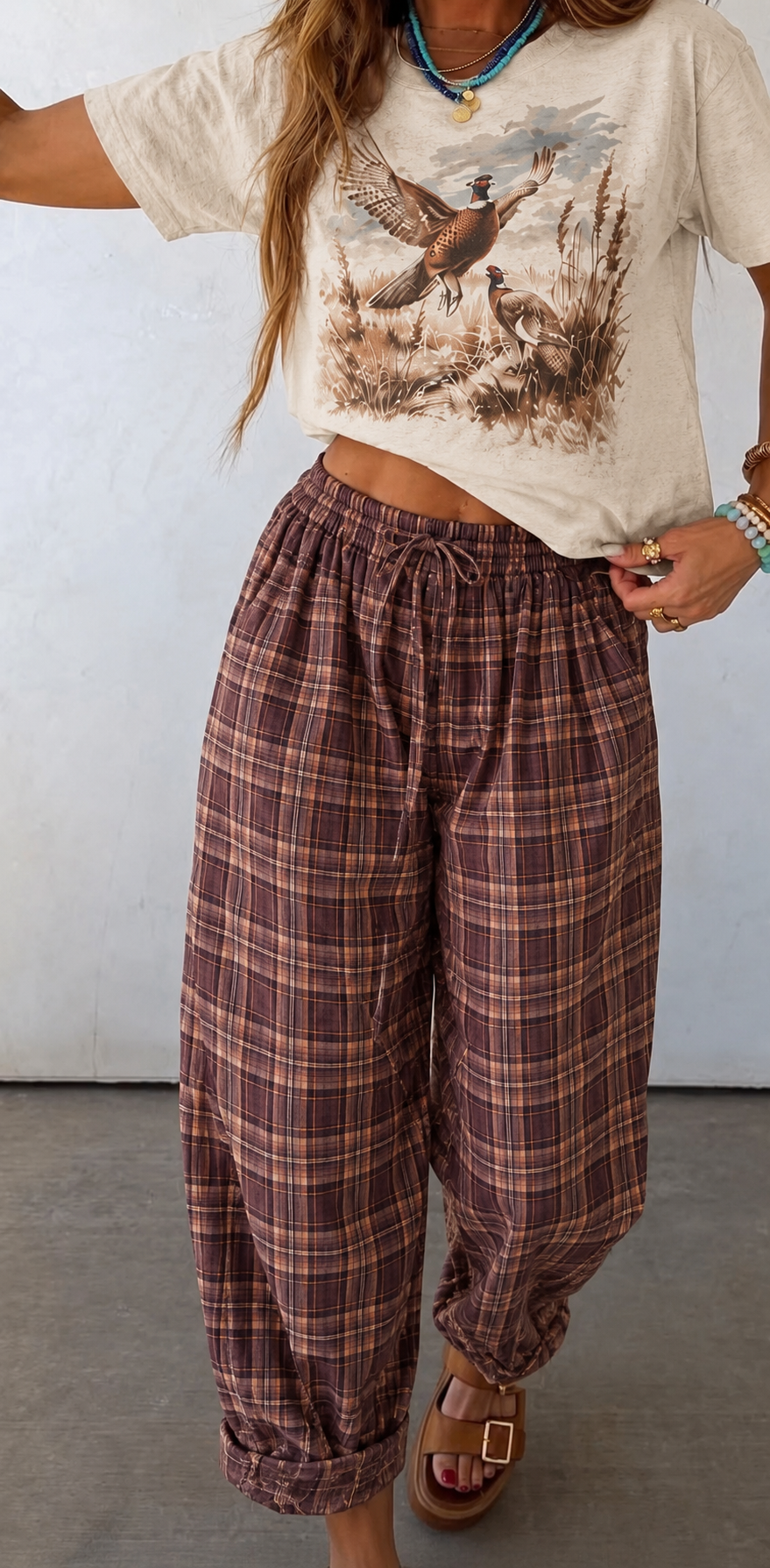 A person wearing a cream graphic T-shirt and loose brown-and-rose plaid drawstring pants, standing against a plain light wall.