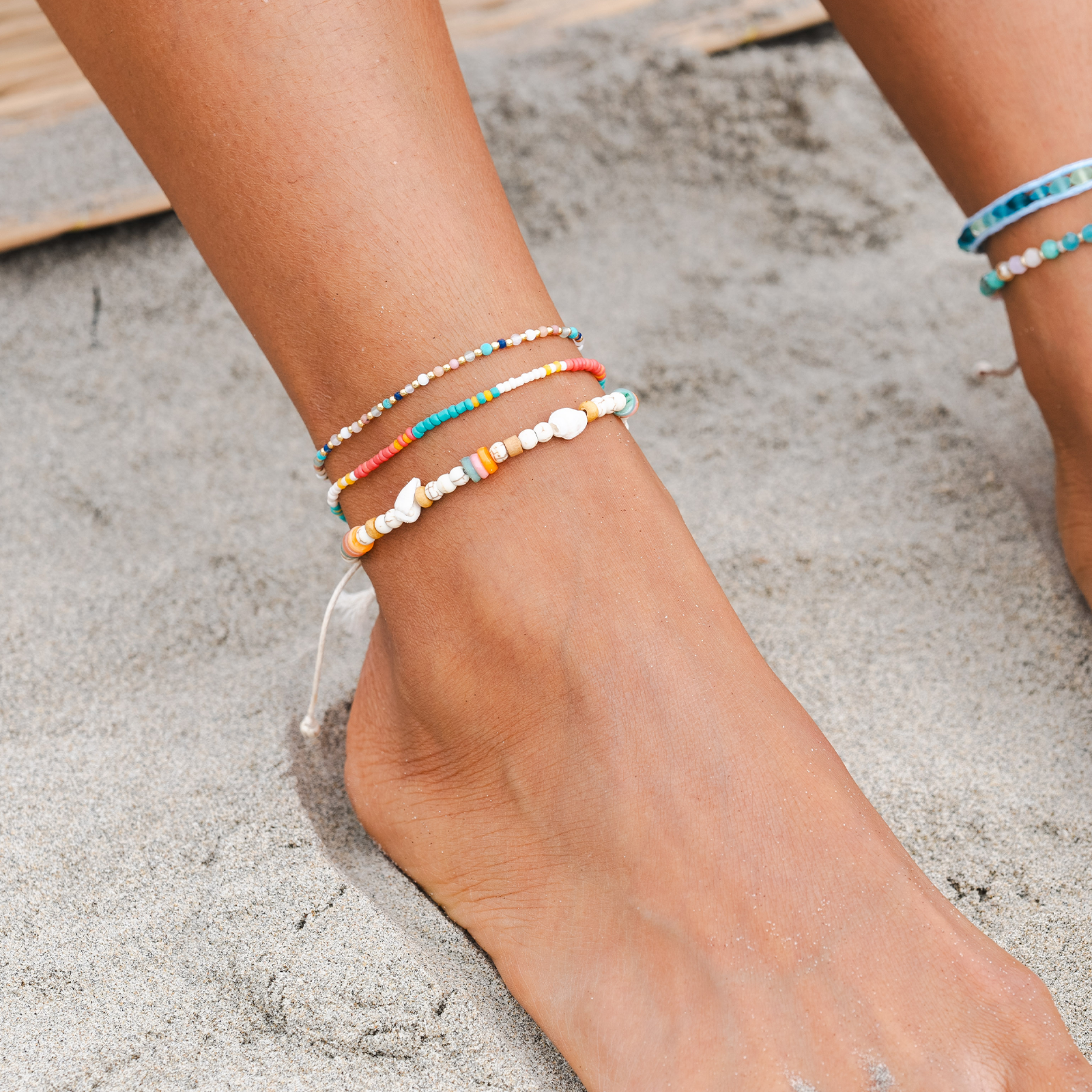 Colorful beaded anklets on a foot against a sandy background