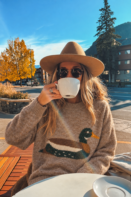 A woman wearing a wide-brimmed tan hat, sunglasses, and a beige knit sweater with a duck motif sits at an outdoor cafe table, holding a white coffee cup. The background features a sunny mountain town scene with autumn trees and a blue sky.